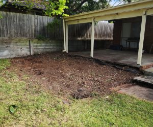 "After photo of a backyard garden bed clearance beside a covered patio. The rectangular bed has been completely stripped of old mulch, plants, and weeds, revealing bare dark soil with scattered twigs and a few emerging sprouts. A concrete patio with white posts, wooden fence, retaining wall, and overhanging tree branches are visible under a partly cloudy sky."